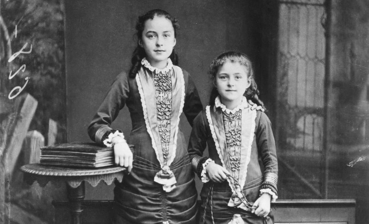 A black-and-white studio photograph of two girls in dark frilled dresses standing side by side, the elder resting a hand on a stack of books on a small table, the younger holding a skipping rope.