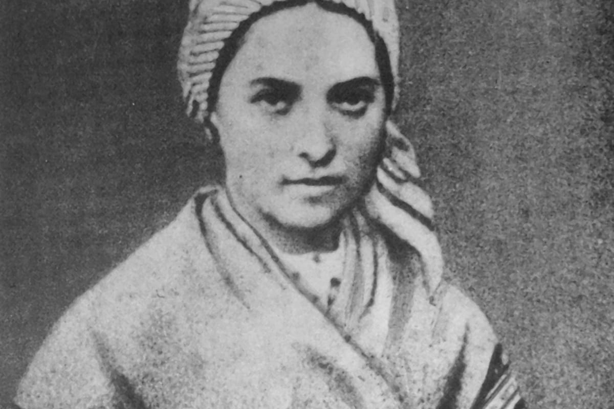 A sepia-toned 1861 photographic portrait of a young Bernadette Soubirous, seated, wearing a plain dress and a headscarf tied under her chin, with a calm, direct expression.