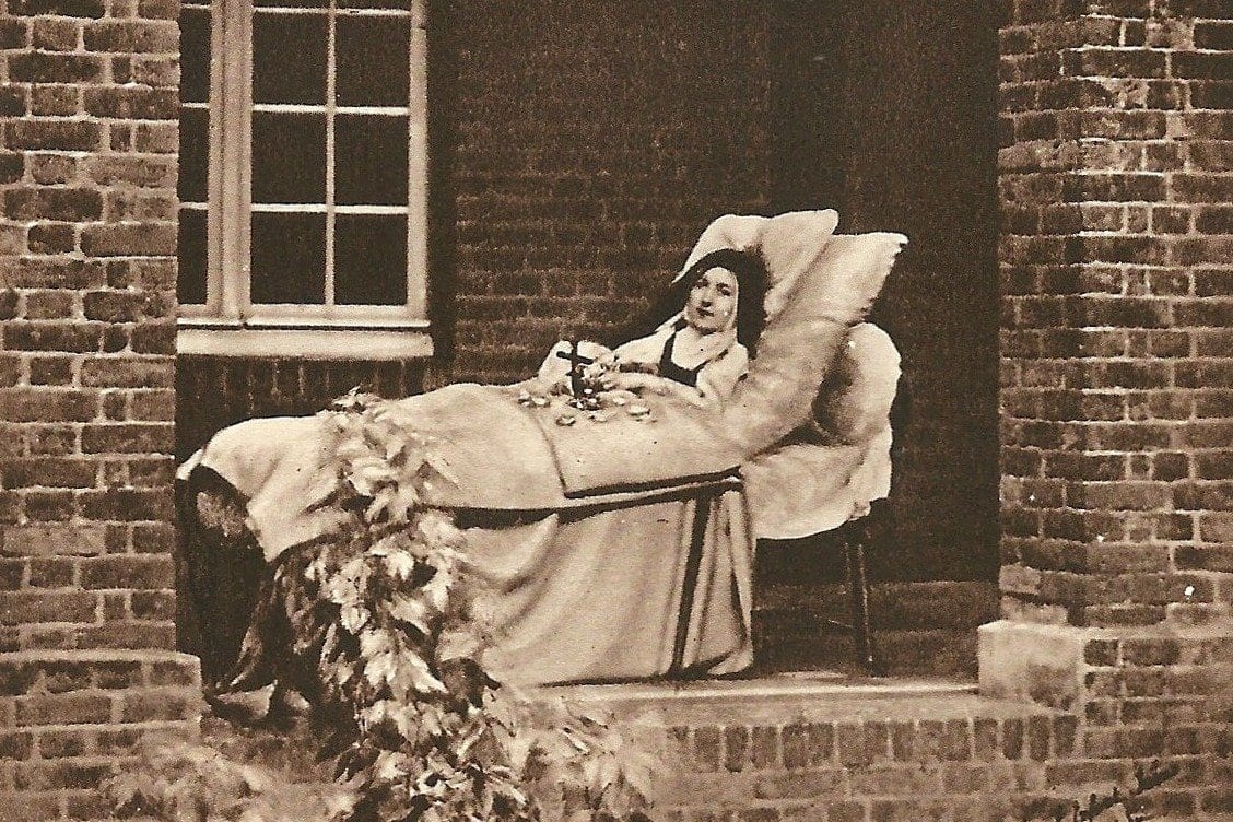 A black-and-white photograph of a young Carmelite nun, gravely ill, propped up on pillows on a bed beneath a stone cloister arcade, her hands holding rose petals.