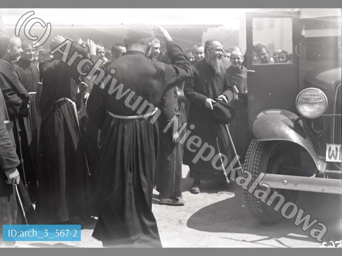 A black-and-white photograph of a bearded friar in a habit, hat in hand, greeted by a crowd of Franciscan friars and boys beside a car parked outside the friary.