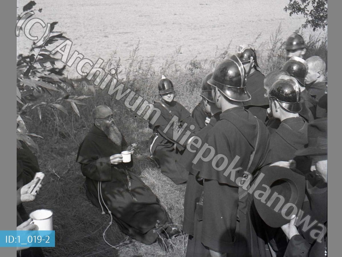 A black-and-white photograph of a bearded, bespectacled Franciscan friar in a dark habit, seated on the grass and holding an enamel cup, surrounded by a group of firemen in metal helmets and dark uniforms, with a field and trees in the background.