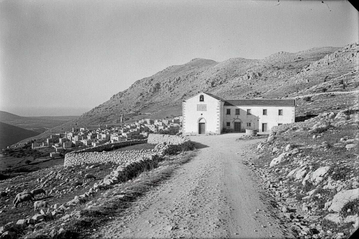 A black-and-white photograph looking up a stony unmetalled mountain road to a small whitewashed church with a bell above its door and a two-storey convent block beside it, with a low stone-walled town on the bare limestone slope below and the sea on the far horizon.
