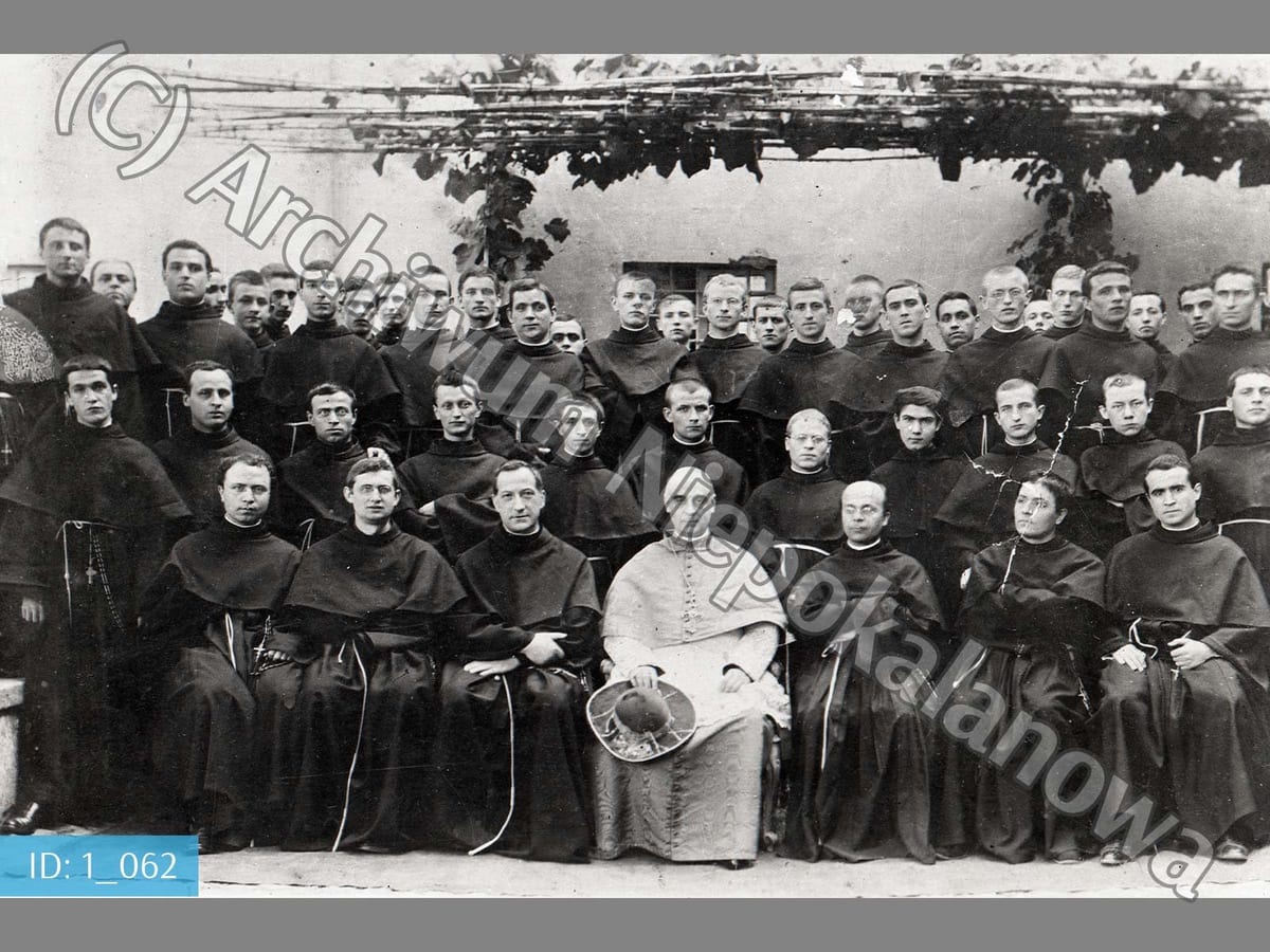 A black-and-white group photograph of several dozen Conventual Franciscan friars in habits, seated and standing in rows under a vine-covered pergola, with a seated prelate in white choir dress at the center of the front row.