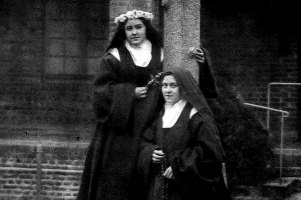 A black-and-white photograph of two Carmelite nuns in habit standing together at the foot of a stone cross in a convent courtyard; the one behind wears a flower wreath over her veil, and the one in front holds a rosary.