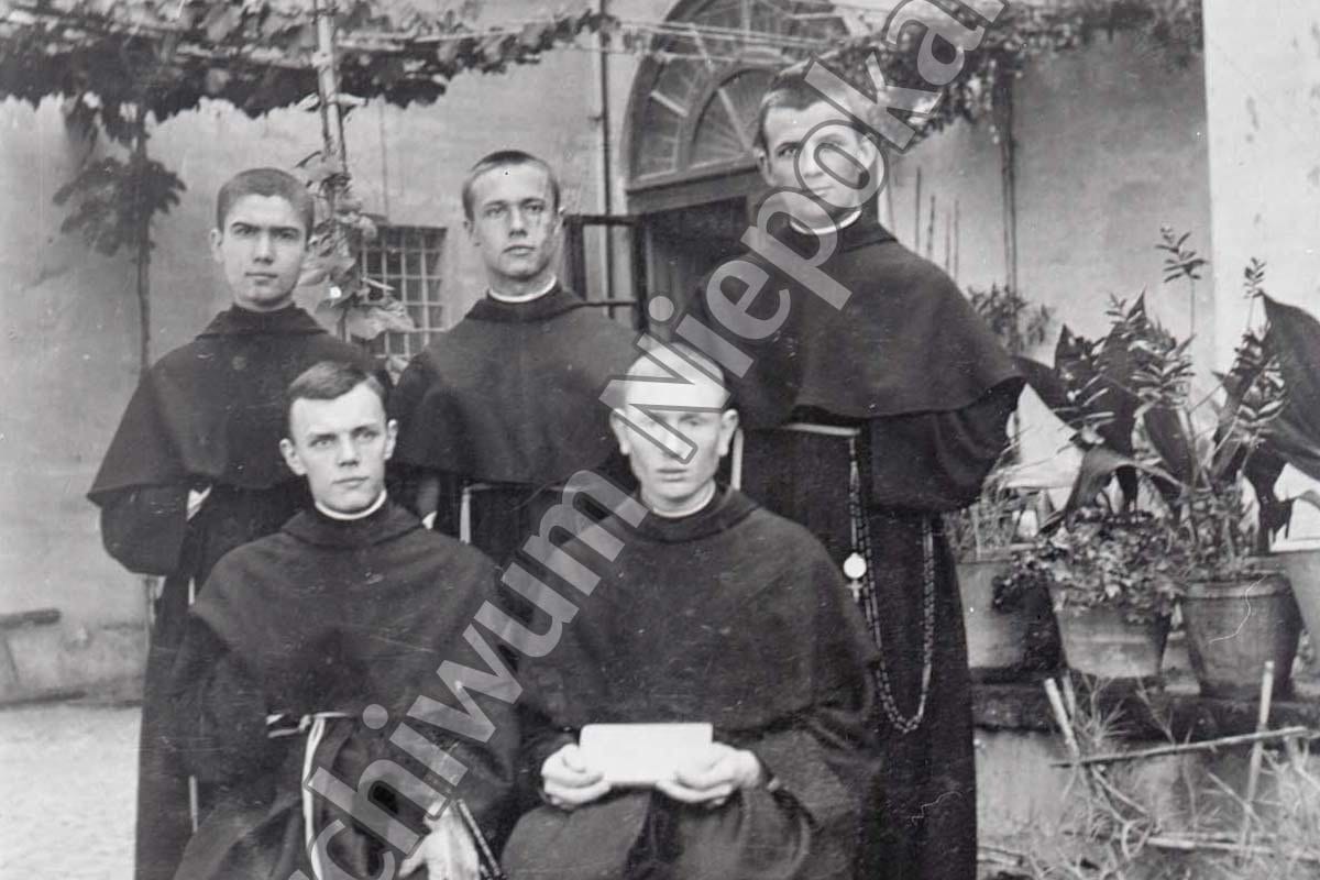 A faded black-and-white photograph of five young men in dark Franciscan habits with rosaries, three standing and two seated, posed in a courtyard before an arched doorway and potted plants; the seated man at center holds a small folded paper.
