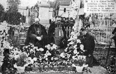 A black-and-white photograph of a bishop and two priests in cassocks standing with bowed heads at the flower-banked grave of St. Thérèse of Lisieux in a cemetery, an iron gate and trees behind them.
