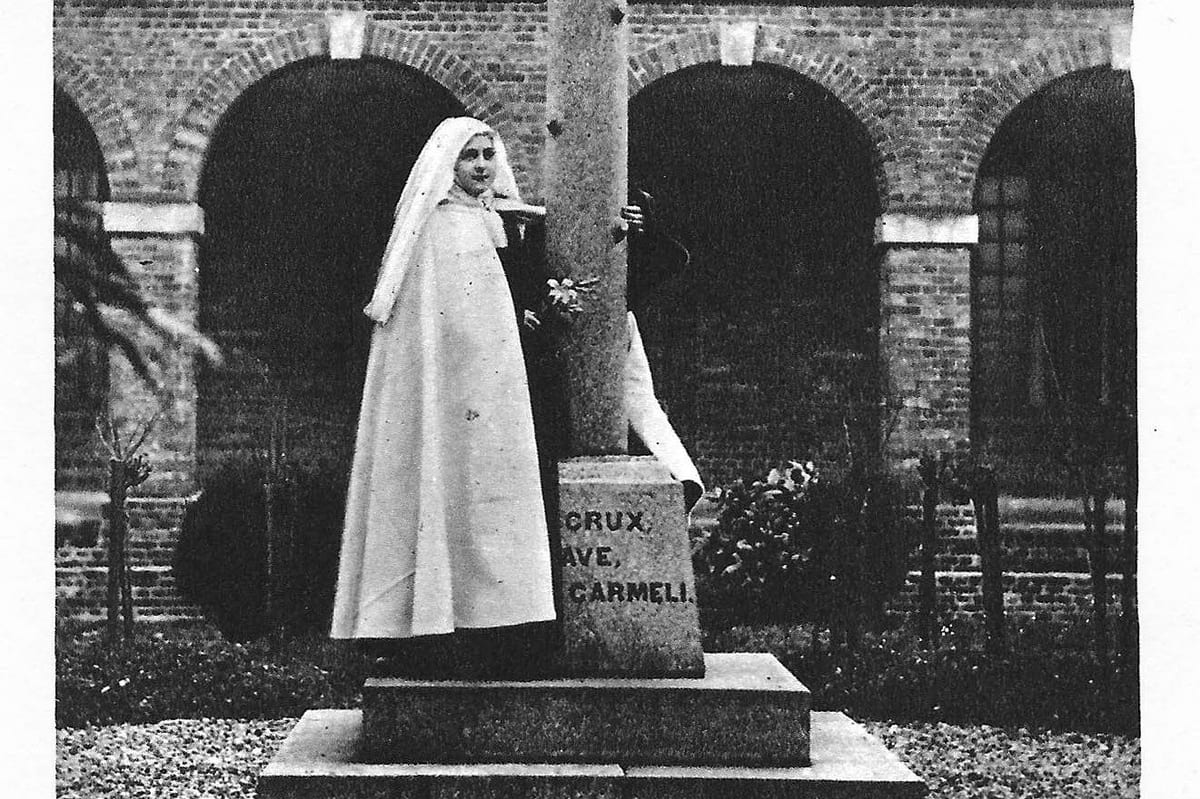 A black-and-white photograph of a young nun in a white novice's habit standing beside a tall stone cross inscribed CRUX, AVE, CARMELI, with a brick cloister arcade behind her.