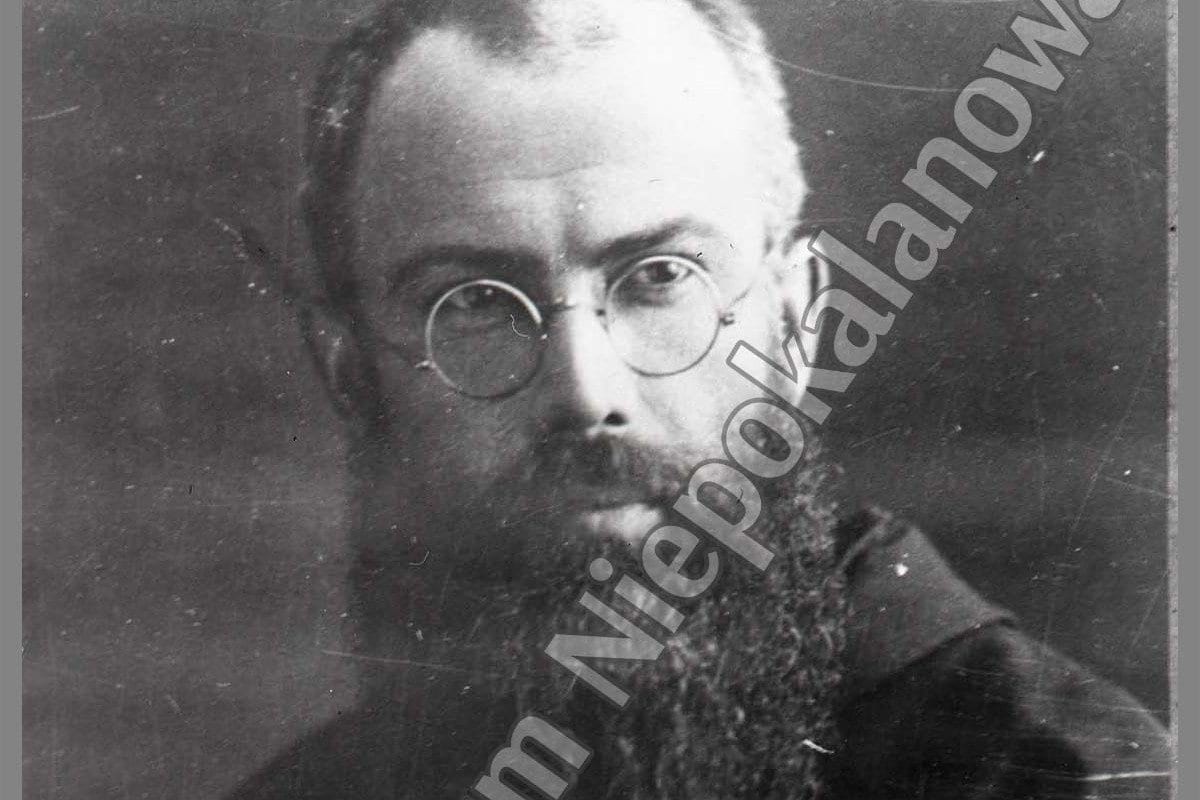 A black-and-white portrait photograph of a bearded Franciscan friar wearing round wire-rimmed glasses and a dark habit, looking directly at the camera.