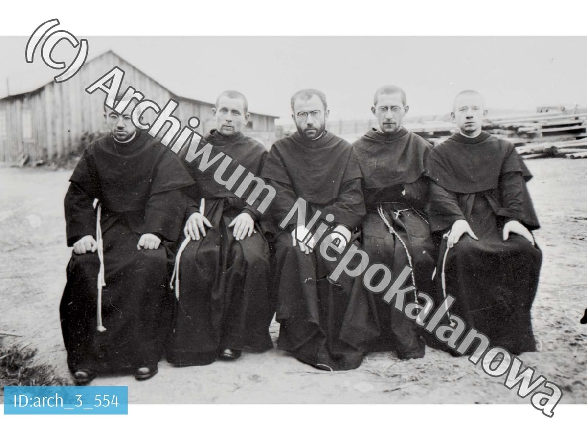 A black-and-white photograph of five Franciscan friars in dark habits, seated outdoors in front of a wooden building; St. Maximilian Kolbe sits in the center wearing glasses and a beard.