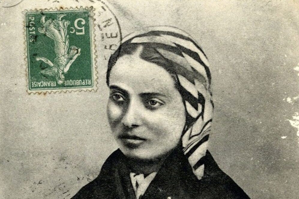 A young woman in a striped headscarf and dark shawl, seated with one hand resting in her lap, looks slightly to the side in a sepia-toned photograph.