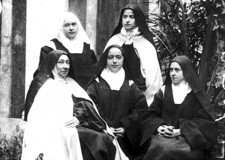 A black-and-white photograph of five Carmelite nuns in habit and veil, three seated and two standing, grouped together in a courtyard with a stone pillar and foliage behind them.