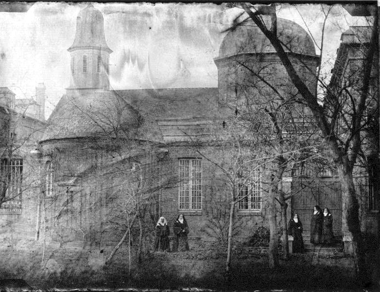 A faded nineteenth-century photograph of a Carmelite monastery building with a domed apse and bare winter trees; two nuns in habits stand at left near the trees, and three more stand together by a doorway and steps at right.