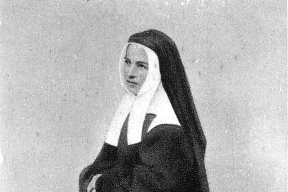 A black-and-white photograph of a young nun in a dark habit and white wimple, seated and turned slightly to one side.