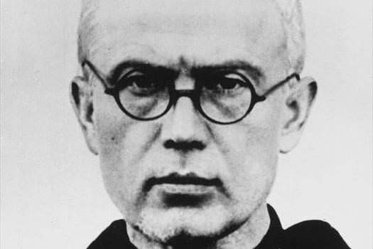 A black-and-white portrait photograph of a bald, bespectacled Franciscan priest in a dark habit, looking directly at the camera with a serious expression.