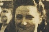 Sepia photograph of a smiling young woman with dark hair pinned up under a headband, in a dark jacket, with a figure partly visible behind her.