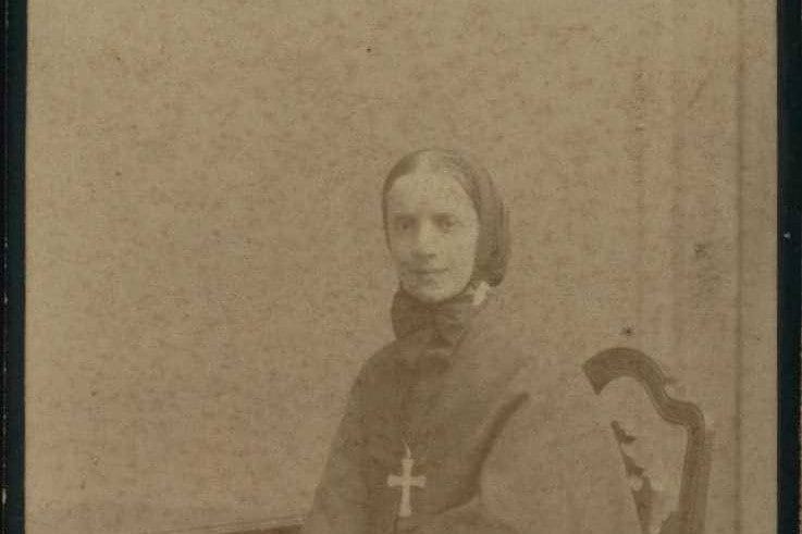 Sepia studio portrait of a young woman in a dark veil, bow and cape with a large cross on her breast, seated beside a carved chair.
