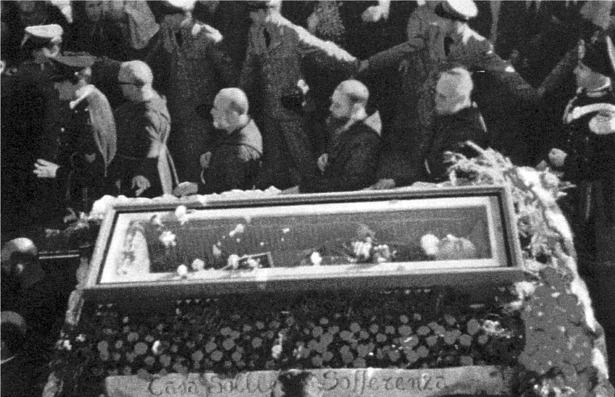 Black-and-white photograph looking down on a glass-topped coffin covered in flowers, with bareheaded Capuchin friars in brown habits and uniformed police standing behind it in a crowd.