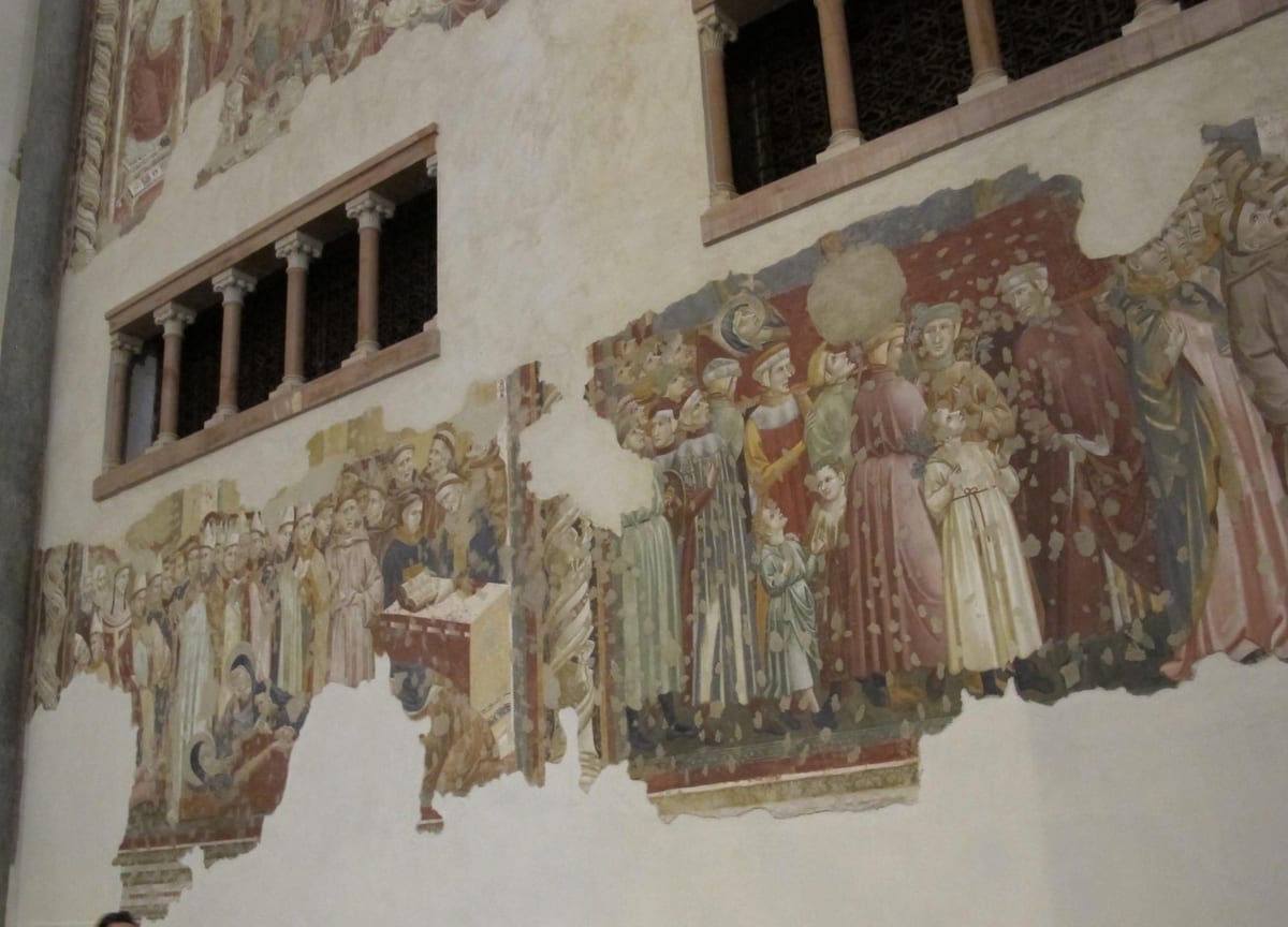 A damaged fresco on a plaster wall: at left, bishops in mitres and friars stand around a lectern with an open book beside the body of St. Clare; at right, a crowd of men and children in red, green and cream robes walks in procession.