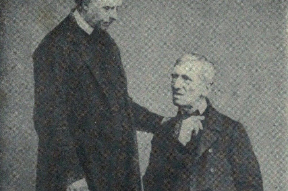 Black-and-white studio photograph of an elderly, grey-haired St. John Henry Newman seated in a dark coat, looking up at a taller priest in clerical collar who stands beside him with a hand on his shoulder.