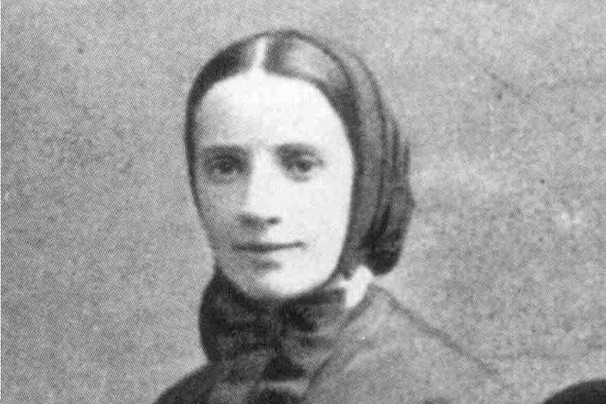 A black-and-white studio photograph of a young nun in a dark habit and headscarf, wearing a large cross on her chest, looking calmly at the camera.