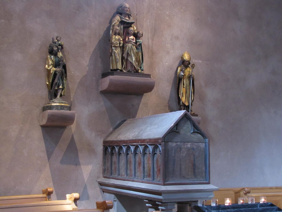A carved and gilded wooden reliquary shaped like a small chapel, standing on a stone pedestal, with a group of painted wooden statues on a shelf above it showing a woman holding a book flanked by three smaller figures, alongside two other statues of male saints, against a bare plaster wall.