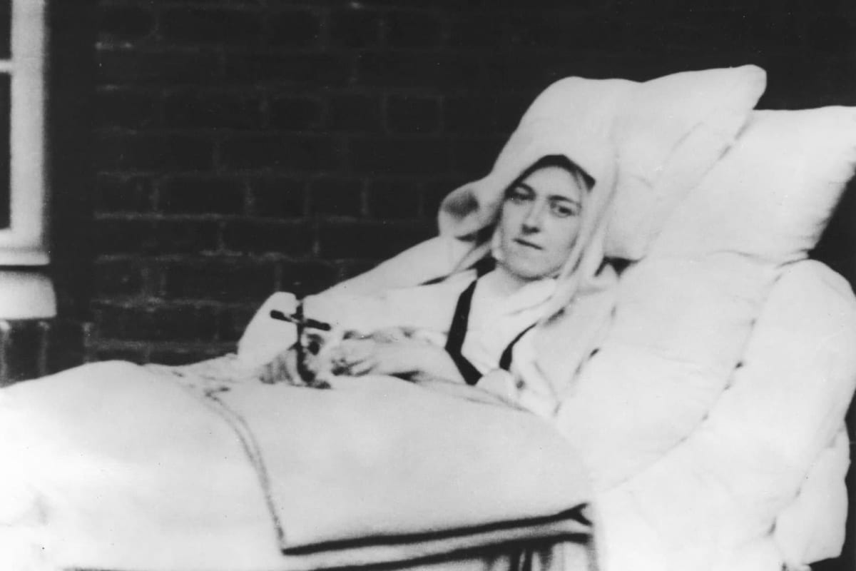 A black-and-white photograph of a young nun lying dead in a white bed, propped on tall pillows, a dark veil framing her face and a small crucifix in her hands, against a brick wall.