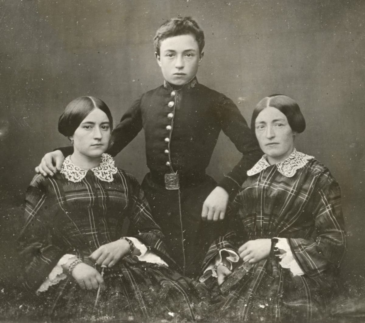 A sepia-toned 1850s studio photograph of two young women in plaid dresses with lace collars seated on either side of a young man in a cadet's uniform standing between them with his arms resting on their shoulders.