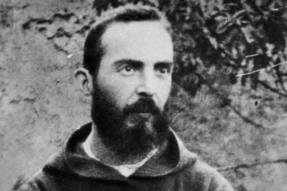 A black-and-white photograph of a bearded young Capuchin friar in a dark habit, standing with arms crossed, a wound visible on the back of each hand, against a wall and foliage.
