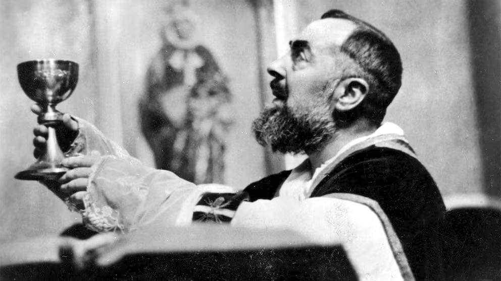 A black-and-white photograph of a bearded Capuchin friar in vestments, bare-handed, raising a chalice above his head during Mass, with a statue visible behind him.