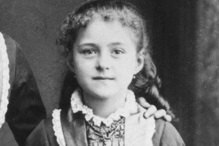 Black-and-white studio photograph of a young girl with long braided hair and a lace-trimmed dress, standing beside an unseen companion whose arm rests on her shoulder.