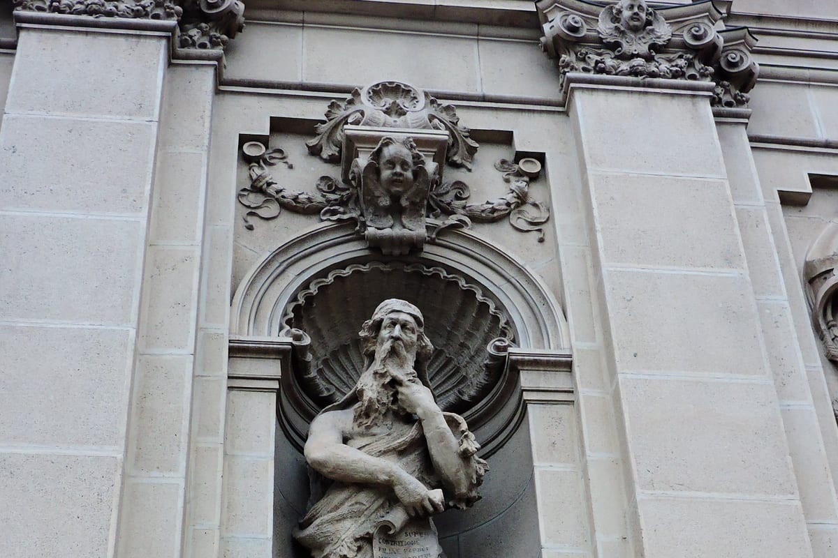 A weathered stone statue of a bearded, robed figure set in a niche on the carved stone facade of Santa Maria Segreta in Milan, beneath an arched shell canopy.