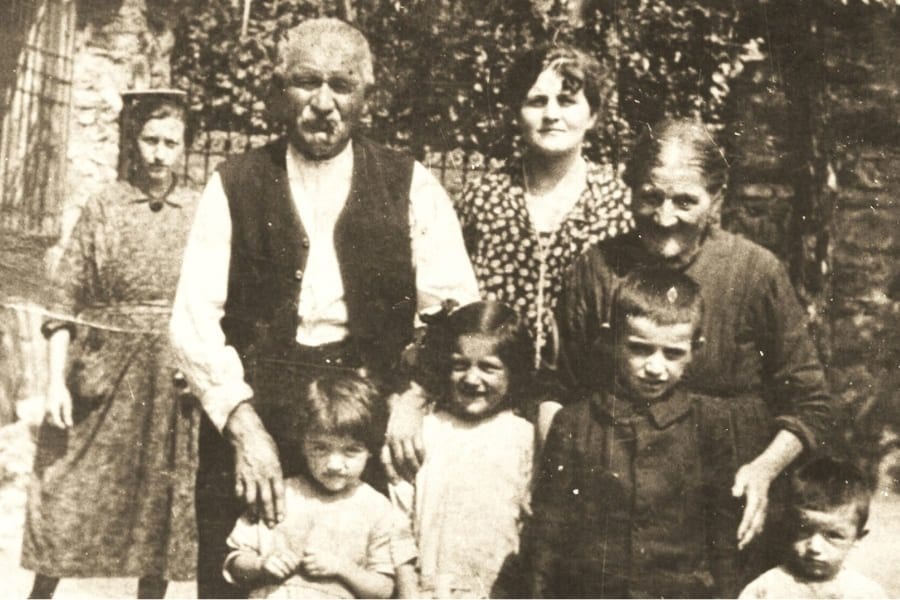 A sepia family photograph: an older man in a dark waistcoat and an older woman stand among a young woman and several children in a garden before a stone wall and foliage.