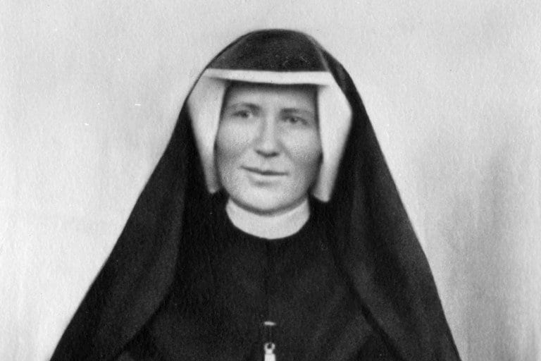 Black-and-white head-and-shoulders portrait of a nun in a dark veil and white wimple, looking calmly toward the camera against a plain pale background.