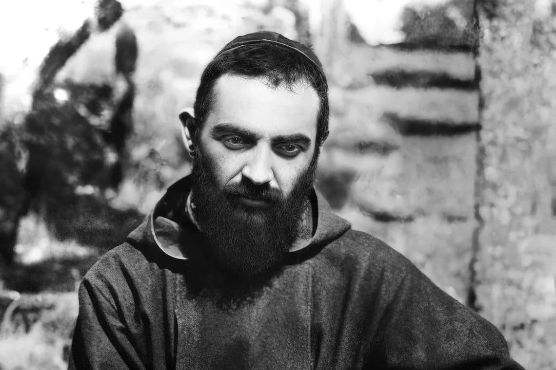 A black-and-white portrait of a bearded friar in a dark hooded habit, looking to the side against a blurred outdoor background.