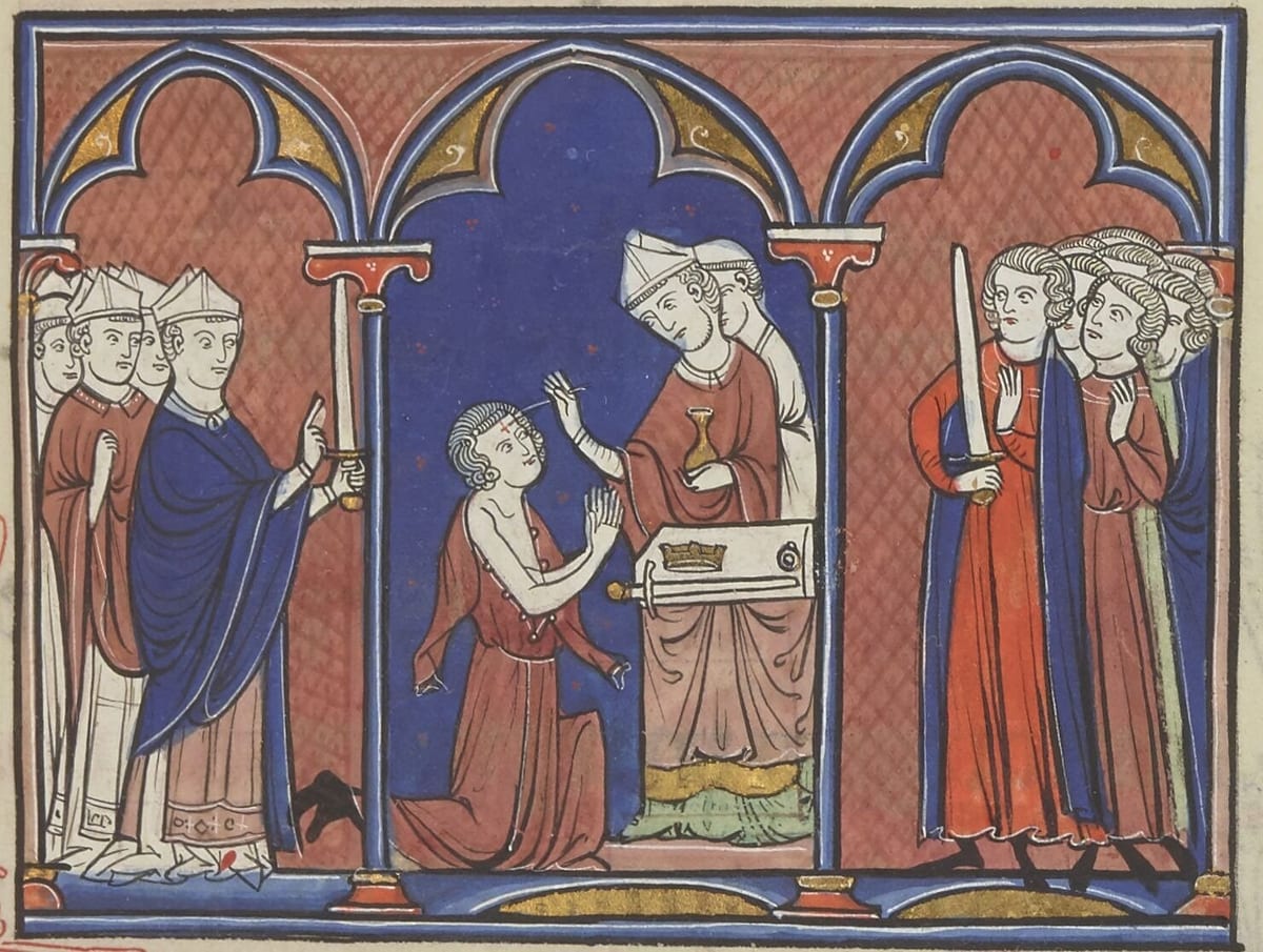 A medieval manuscript painting in blue, red and gold: a bishop in a white mitre anoints the forehead of a kneeling, bare-shouldered boy with oil from a flask, beside an altar bearing a crown, while mitred clergy stand at the left and armed lords watch from the right.