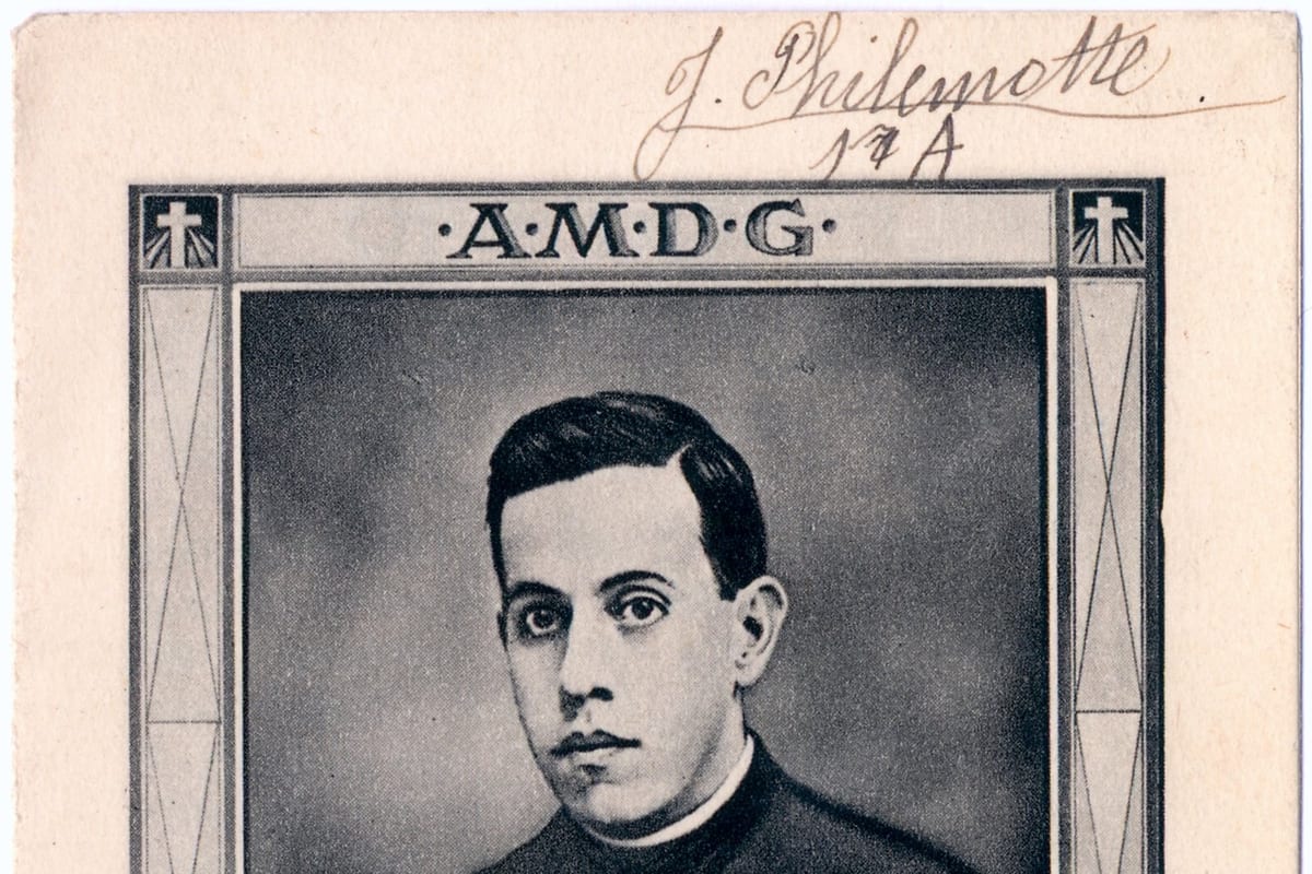 Black-and-white portrait of a young man in a black cassock and white collar, his head turned slightly, framed within a holy card bearing the letters AMDG.