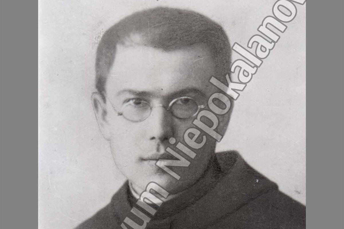 Black-and-white studio portrait of a young man with short dark hair and round wire-rimmed glasses, wearing a dark high-collared habit against a pale background.