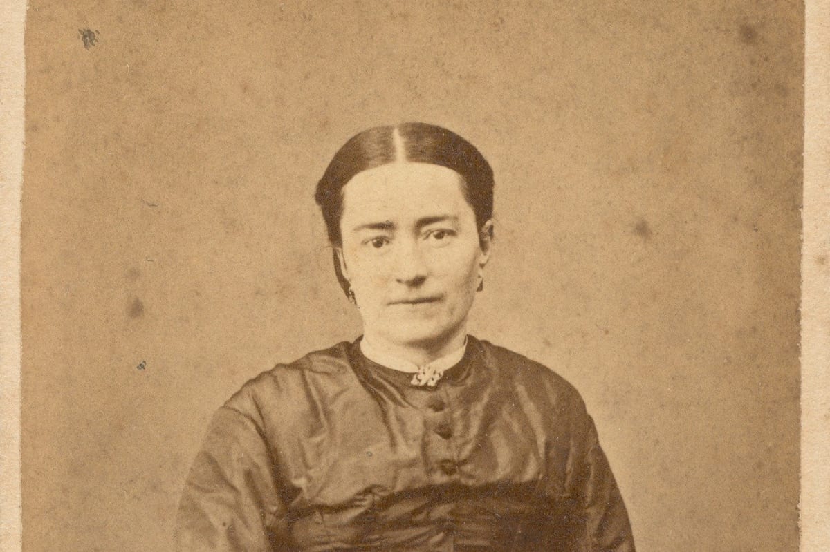 A sepia-toned 19th-century studio photograph of a woman with dark hair parted in the center, wearing a dark silk dress buttoned to the neck, seated with her hands folded in her lap.