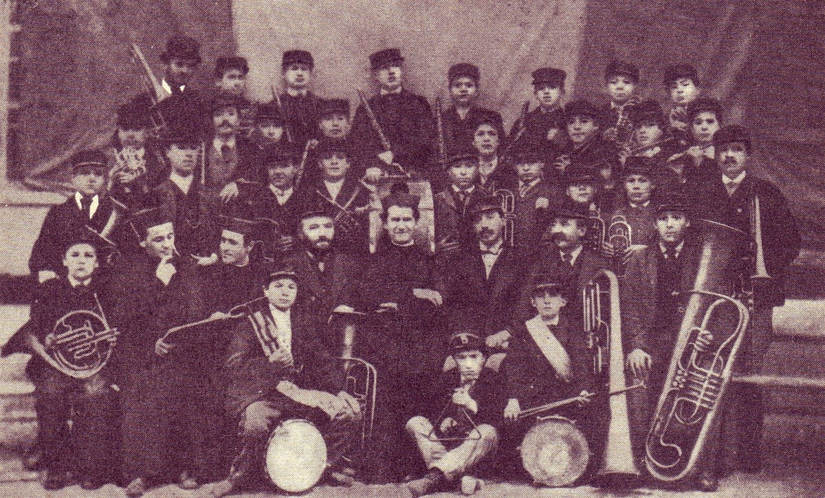 A sepia-toned archival photograph of St. John Bosco standing among dozens of boys outside a building, many holding brass instruments and drums.