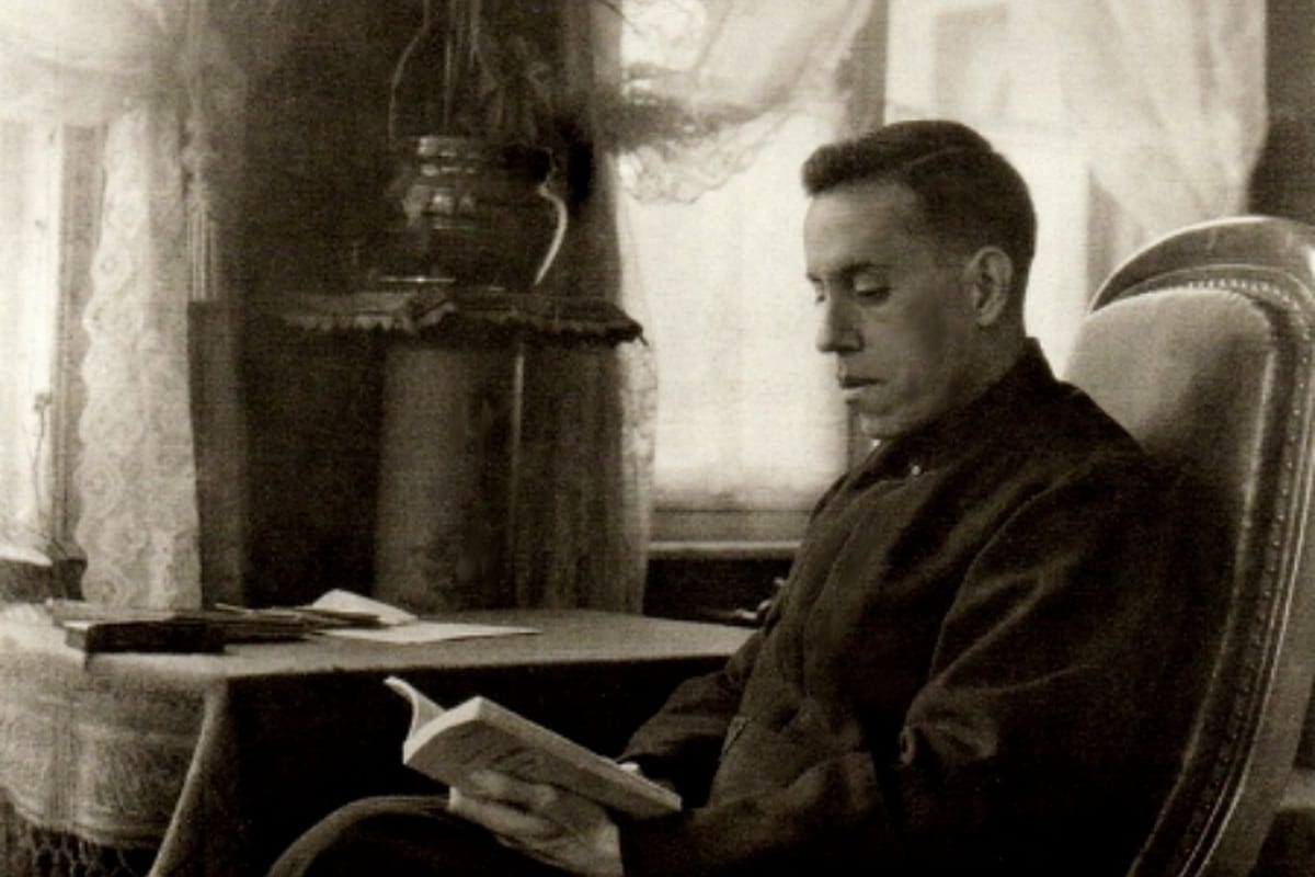 A black-and-white photograph of a young man in a dark coat seated in an armchair beside a writing table, reading from an open book, with lace curtains and a stove behind him.
