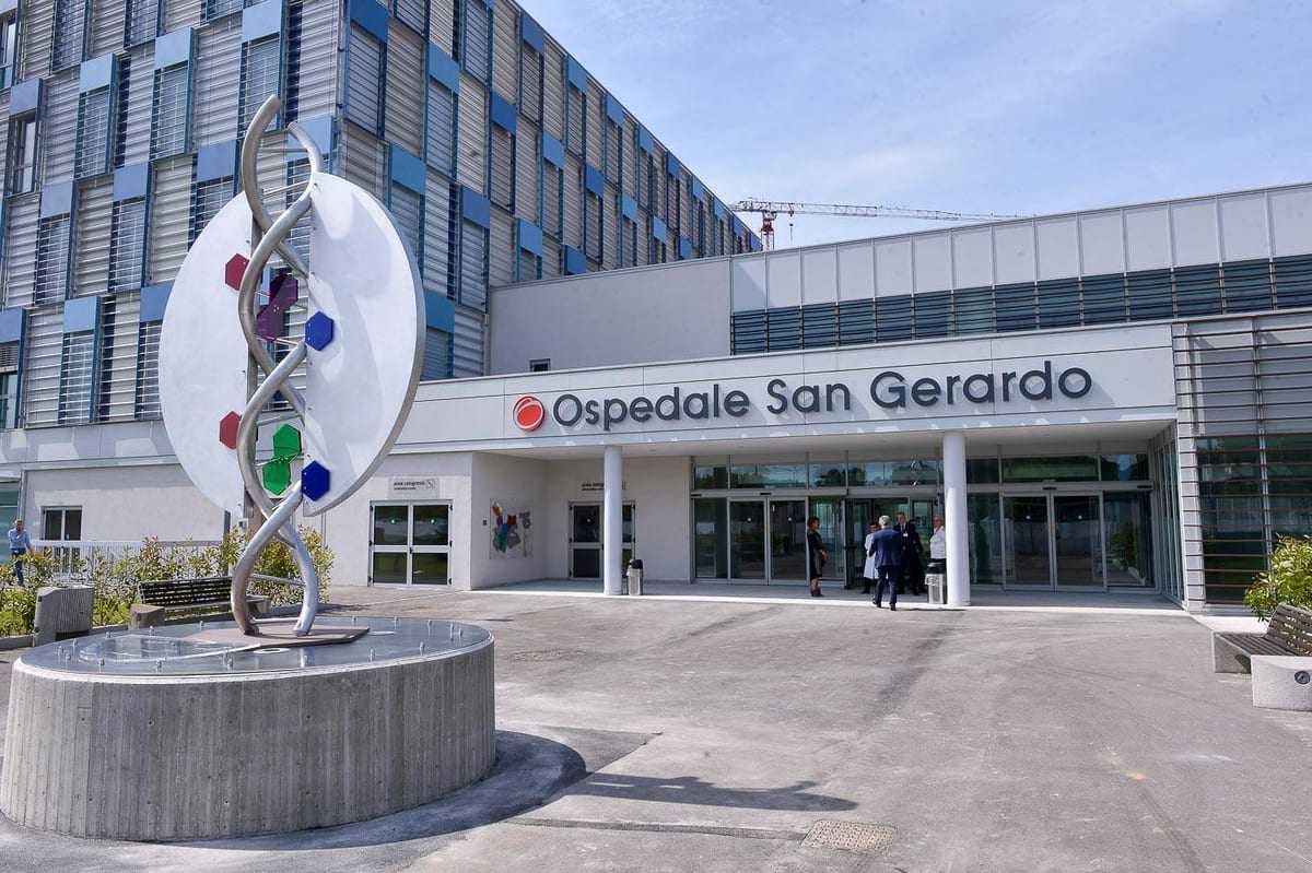 The glass-fronted entrance of Ospedale San Gerardo in Monza, with a DNA-helix sculpture in the foreground and visitors walking toward the doors.