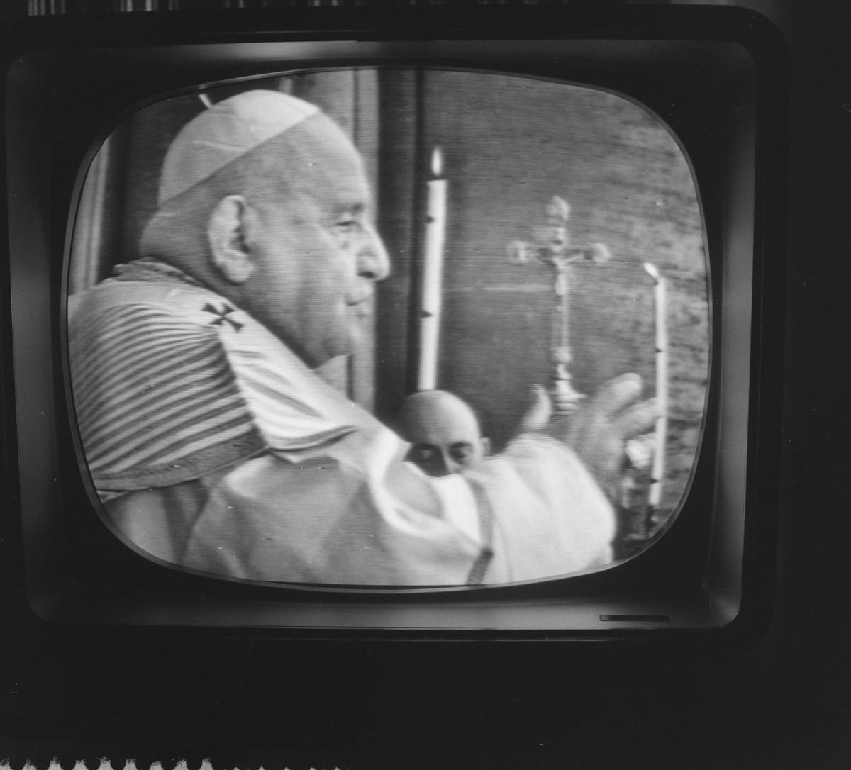 A black-and-white photograph, shown on a television screen, of Pope John XXIII in profile, wearing a white cassock and skullcap, raising his hand in blessing before a crucifix and candles.