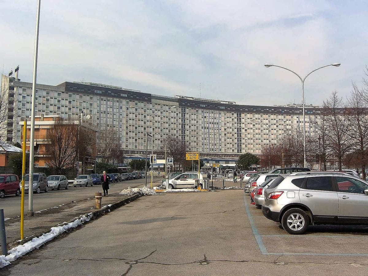 A wide, multi-storey hospital building seen across a parking area on a winter day, with bare trees, parked cars and patches of snow in the foreground.