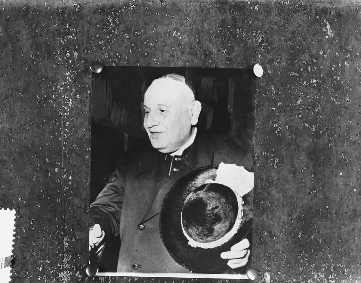 A black-and-white photograph of an elderly cardinal in clerical dress, smiling slightly and holding a wide-brimmed hat in both hands.
