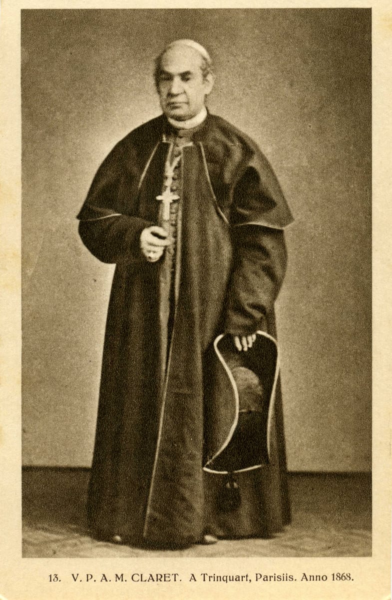 A photograph of the archbishop standing full length in a Paris studio, in cassock and caped cloak, holding his pectoral cross.