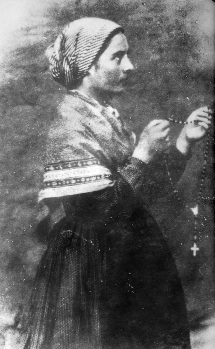 A young woman in a headscarf and shawl, seen in profile, holds a rosary raised in her clasped hands, in an 1861 AD photograph taken at Lourdes.