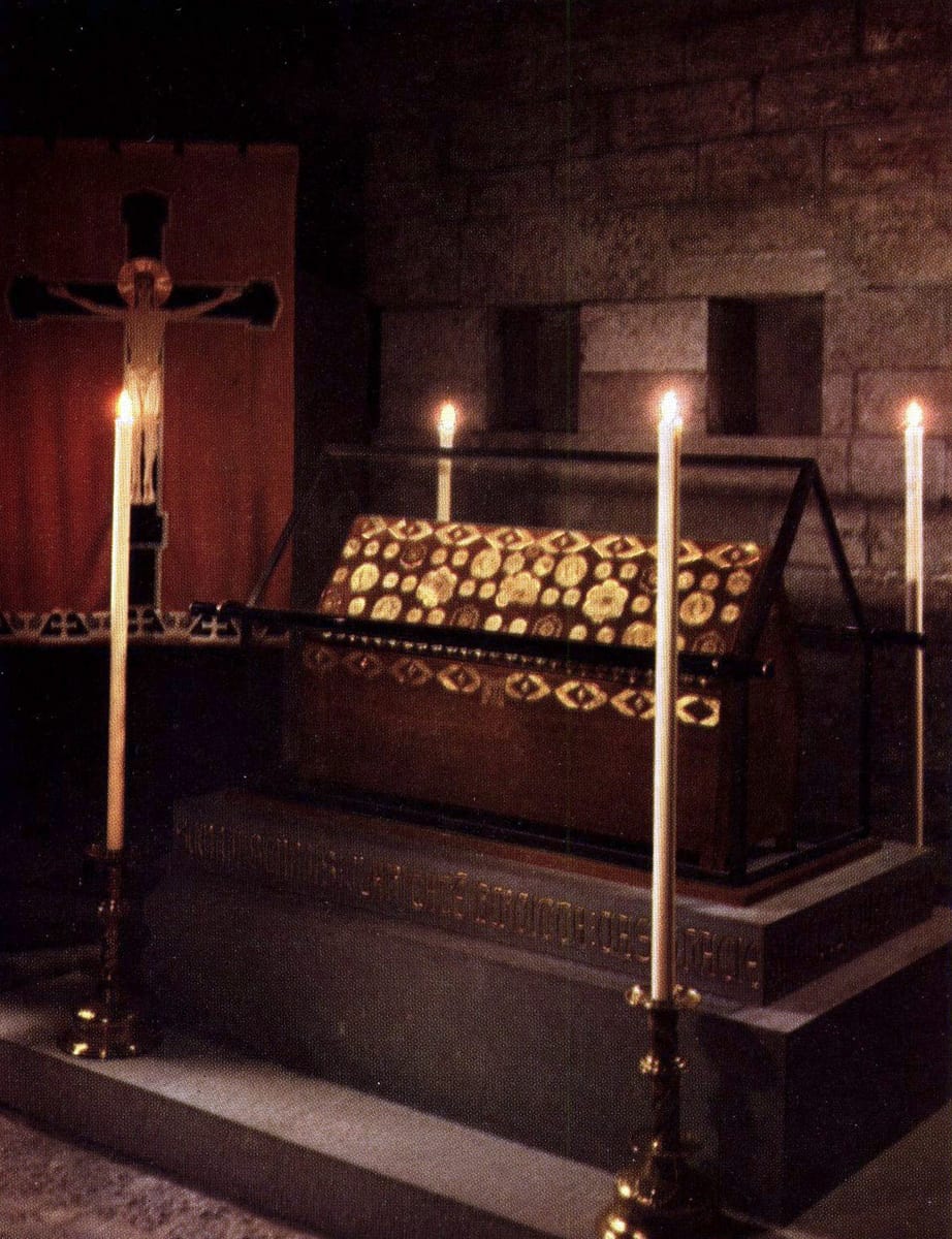 A candlelit photograph shows a gabled reliquary shrine covered in dark red velvet with gilded roundels, standing in a glass case with tall candles around it.