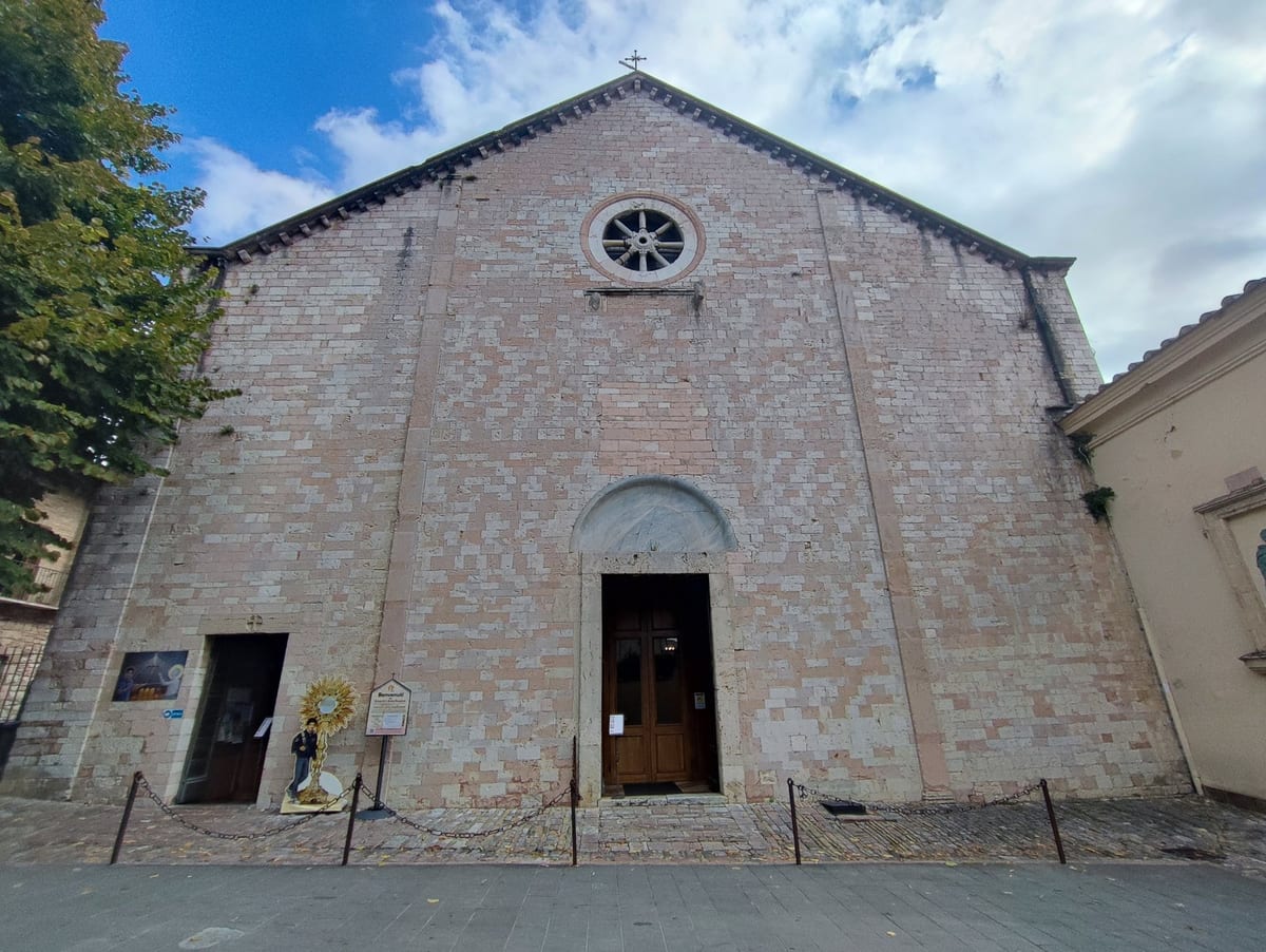 The pale pink stone front of the church of Santa Maria Maggiore in Assisi, with a round rose window above an arched wooden door, and a standing cut-out figure of a boy beside a golden monstrance to the left of the entrance.