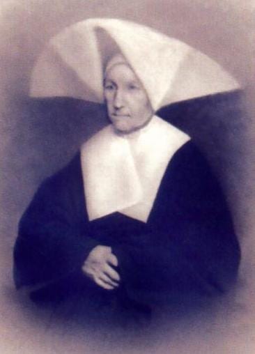 An elderly woman in the winged white cornette and dark habit of the Daughters of Charity sits with her hands folded in her lap, photographed in 1876 AD.