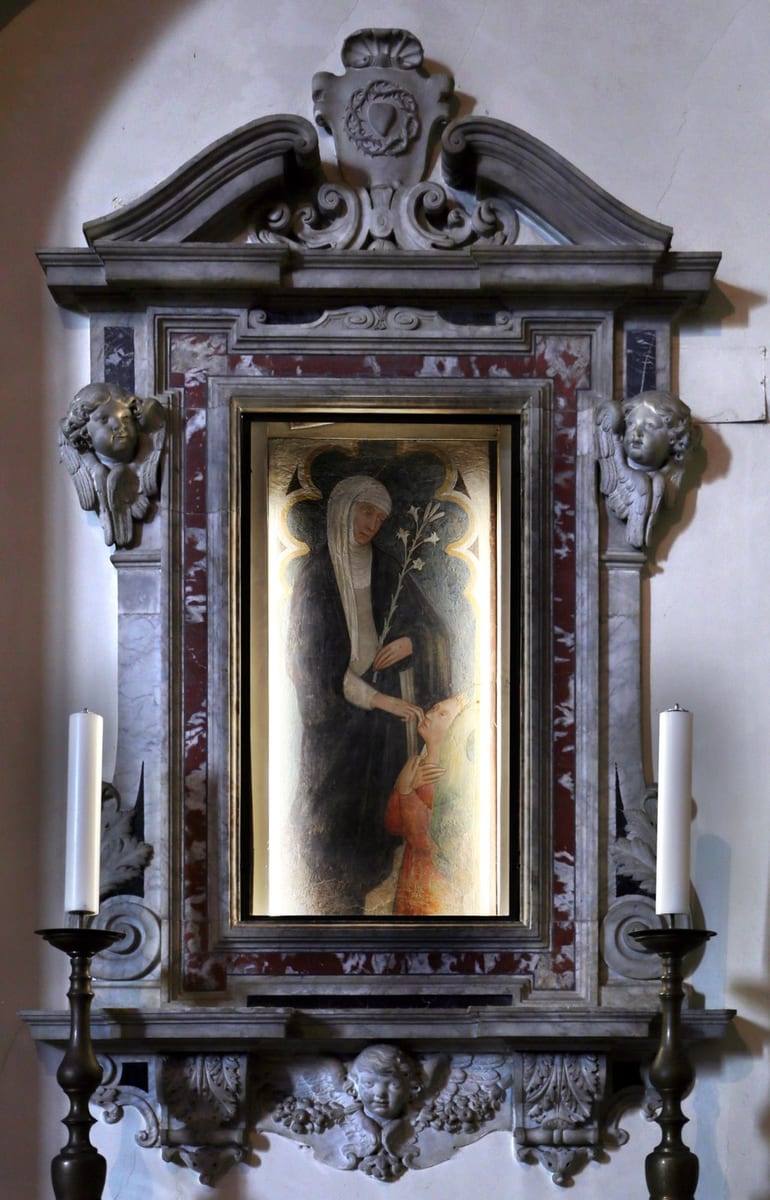 A fresco of Saint Catherine of Siena in a black-and-white Dominican habit, holding a lily and touching the chin of a kneeling figure in red before her, set within an ornate marble altar frame flanked by carved angel heads and candles.